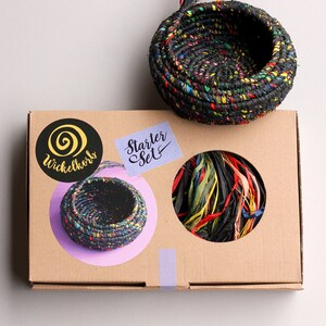 Peut inclure: Une boîte en carton contenant un panier tissé noir et coloré. La boîte a un autocollant qui dit "Starter Set" et un autre autocollant avec un logo qui dit "Wickelkorb".