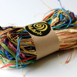 Può includere: Un rotolo di carta triturata colorata, perfetto per confezionare regali o per lavori artigianali. La carta è un mix di marrone naturale e colori vivaci come blu, verde, rosso e giallo. Il rotolo è avvolto in carta marrone con un'etichetta nera su cui è scritto "Wichtelskorb" e ha un design a spirale giallo.