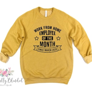 May include: Mustard yellow sweatshirt with black text that reads "WORK FROM HOME EMPLOYEE OF THE MONTH SINCE MARCH 2020". The sweatshirt has a crew neck and long sleeves with rolled cuffs. The text is centered on the front of the shirt.
