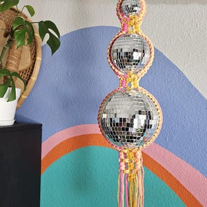 May include: A macrame hanging with three disco ball ornaments. The macrame is made with pink and yellow yarn and has a fringe at the bottom.