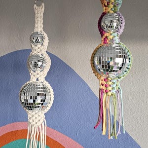 May include: Two hanging disco ball ornaments made of macrame with mirrored balls. The macrame is white and cream on the left and multi-coloured on the right.