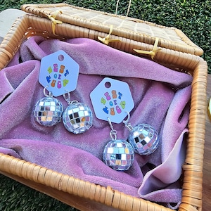 Peut inclure: Deux paires de boucles d'oreilles en forme de boule disco argentée dans un panier en osier doublé de velours rose. Chaque boucle d'oreille est ornée d'un petit charme en forme de boule disco argentée et d'un charme plus grand en forme de boule disco argentée. Les boucles d'oreilles sont étiquetées avec une étiquette blanche qui dit "Best of the Fuckin'"