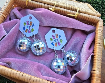 May include: Two pairs of silver disco ball earrings in a wicker basket lined with pink velvet. Each earring features a small silver disco ball charm and a larger silver disco ball charm. The earrings are labeled with a white tag that says "Best of the Fuckin'"