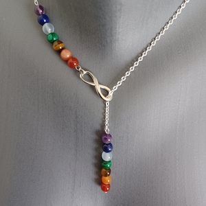 Könnte beinhalten: Silberfarbene Halskette mit einem Anhänger in Form eines Unendlichkeitszeichens. Die Kette hat eine Reihe farbiger Edelsteinperlen in einer vertikalen Linie. Die Perlen umfassen Lila, Blau, Grün, Orange und Rot.