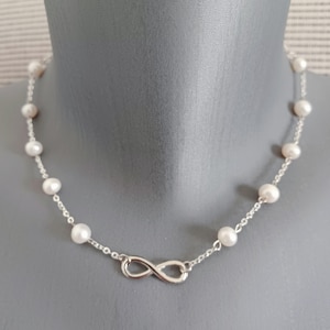 May include: A necklace with white pearls and a silver chain. The necklace has an infinity symbol pendant. The pearls are evenly spaced along the chain, creating a classic and elegant look. The necklace is a timeless piece.