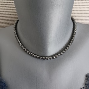 May include: A dark gray beaded necklace displayed on a mannequin. The necklace is composed of small, round beads, creating a simple, elegant design. The beads have a metallic sheen, reflecting light. The necklace is a choker style.