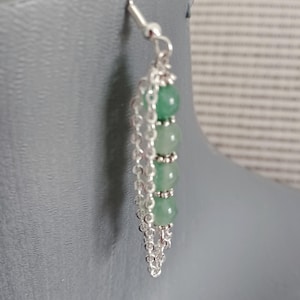 Può includere: Un singolo orecchino con un design a gancio e catena argentati. L'orecchino presenta quattro perline verdi rotonde e una catena d'argento che pende sotto. L'orecchino è esposto su uno sfondo grigio.