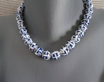 May include: A beaded necklace featuring round, white beads with a blue geometric pattern. The necklace is displayed on a gray mannequin. The beads are strung together to form a necklace.