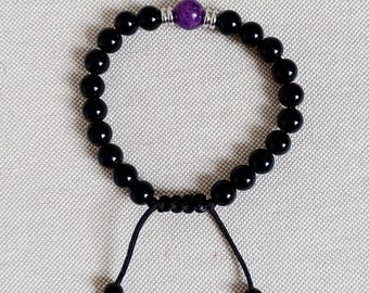 Puede incluir: Pulsera de cuentas negras con una cuenta central morada. La pulsera es ajustable con un cierre de nudo corredizo y cordones negros. Las cuentas son redondas y brillantes. Fondo neutro.