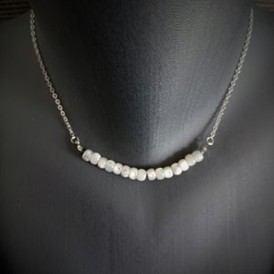 May include: A delicate necklace featuring a silver chain and a row of small, white, rectangular beads. The beads are strung together to form a gentle curve, creating a minimalist and elegant design. The necklace is displayed on a black mannequin.