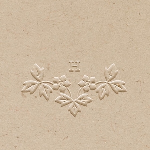 May include: A beige card with an embossed floral design. The design includes three flowers and leaves, with the letter "H" above the flowers. The card has a textured surface.