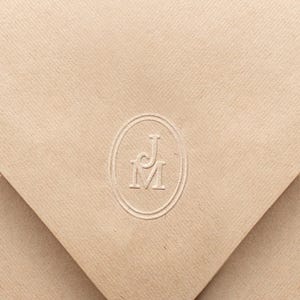 May include: A tan envelope with a textured surface. The envelope is sealed and features a circular embossed design with the letters "JM". The envelope's design includes diagonal lines.