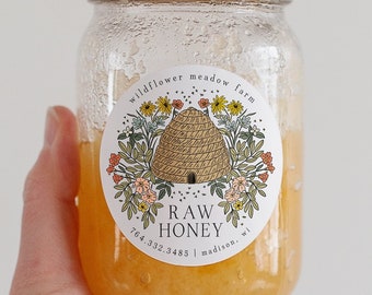 May include: A clear glass jar filled with raw honey, held against a white background. The jar has a gold lid and a label with a beehive illustration and the text "Raw Honey". The honey is a golden yellow color.