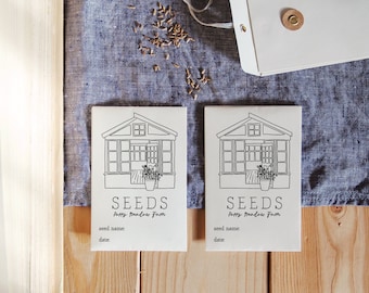 May include: Two white seed packets with black line drawings of a greenhouse and the text "SEEDS Happy Meadow Farm" on a wooden table with a book and a blue linen cloth.