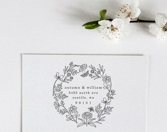 May include: A white card with a black and white floral wreath design. The text inside the wreath reads "autumn & william 5485 north ave seattle, wa 98101"