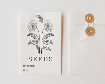 May include: A white card with black ink reads "SEEDS" and features a simple line drawing of two flowers with leaves and a butterfly. The card also has the text "seed name:" and "date:" printed on it. The card is in a white envelope with two gold metal fasteners.
