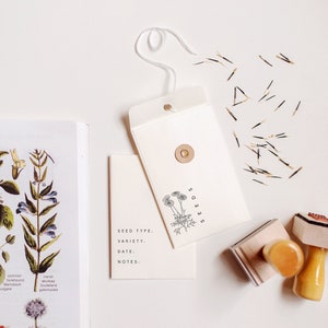 May include: A white envelope with a brown button closure and the word "SEEDS" printed on it. A small white card with the words "SEED TYPE, VARIETY, DATE, NOTES." is next to the envelope. There are also two wooden stamps and a pile of small brown seeds.