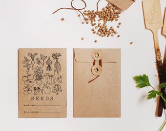 May include: A brown paper envelope with a string closure and a label that says "SEEDS" with a drawing of various vegetables and herbs. The envelope is sitting on a white surface with a small pile of seeds and two wooden plant markers.