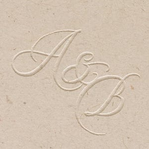 May include: A close-up of a beige paper with embossed initials "A & B" in a delicate, cursive font. The paper has a textured surface and a subtle, neutral color, suitable for stationery or invitations.