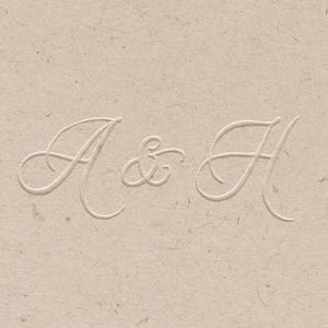 May include: A close-up of a textured, beige paper with embossed lettering. The letters "A & H" are elegantly scripted in a delicate, cursive font. The design is simple and refined, suitable for stationery or invitations.