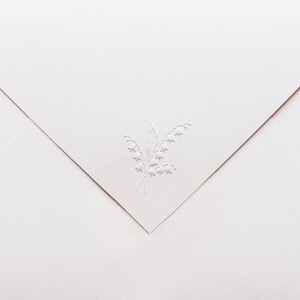May include: A light pink envelope with a detailed embossed lily of the valley design in the upper right corner. The envelope is folded in a classic triangular shape, with the embossed design adding a touch of elegance. The paper has a subtle texture.