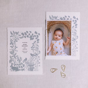 Puede incluir: Una tarjeta blanca con un borde floral azul y el texto "Introducing Peter Benjamin born 05.22.23 8:39 am"  Una foto de un beb&eacute; en una cesta con un mono blanco y naranja.