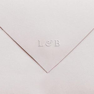 May include: A light pink envelope with the embossed letters "L & B". The envelope is folded at the corner, revealing a triangular shape. The paper has a textured surface. Ideal for wedding invitations or stationery.