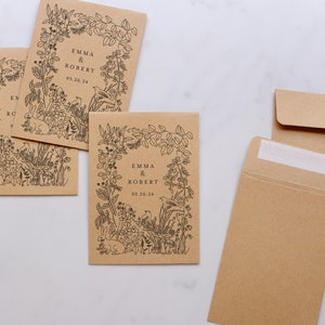 Op de afbeelding: Kraftpapieren zaadzakjes met een zwart bloemenontwerp en de namen "EMMA & ROBERT" en de datum "05.26.24". De zakjes bevatten illustraties van planten, vogels en een konijn. Een open envelop is ook zichtbaar.