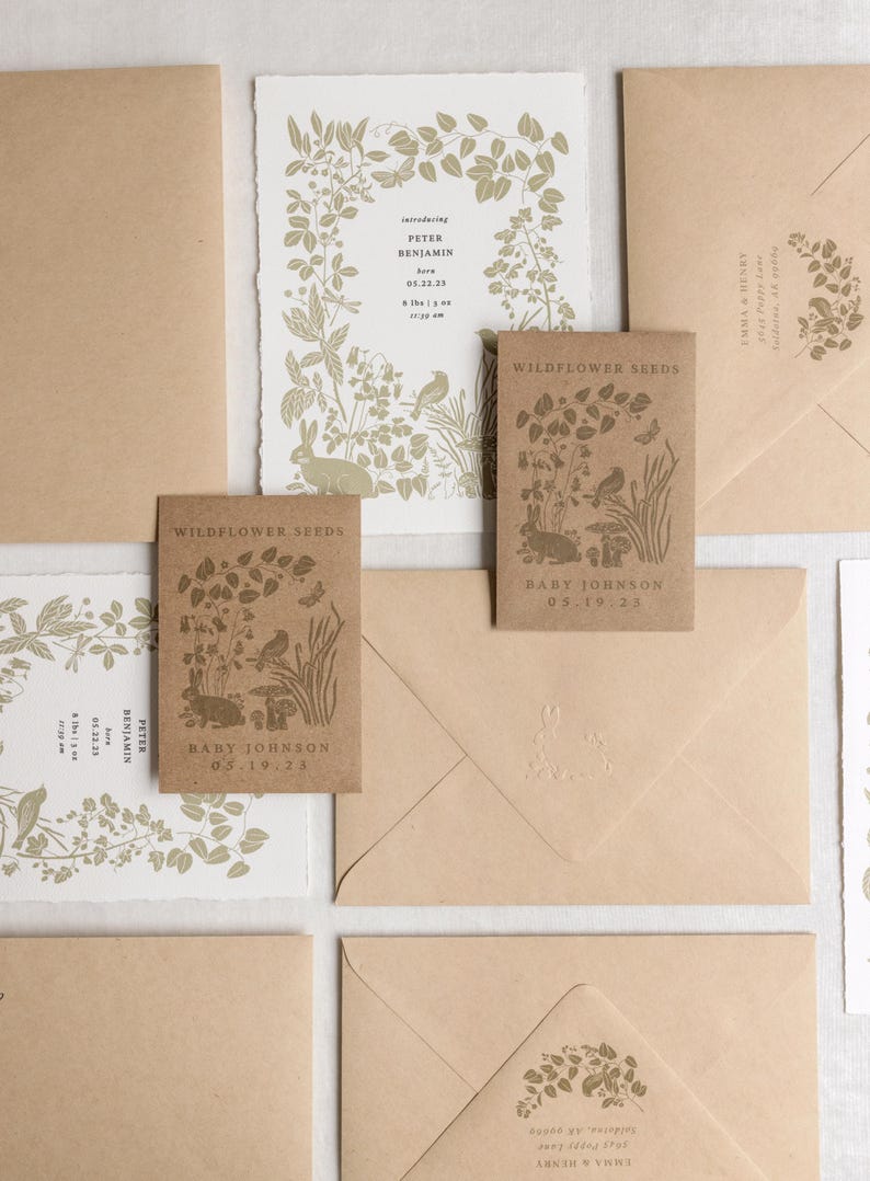 K&ouml;nnte beinhalten: Eine Sammlung von Babykarten und Wildblumensamen-P&auml;ckchen. Die Karten zeigen botanische Illustrationen und Text, einschlie&szlig;lich Babynamen und Geburtsdaten. Die Umschl&auml;ge sind hellbraun und haben ein passendes Design.