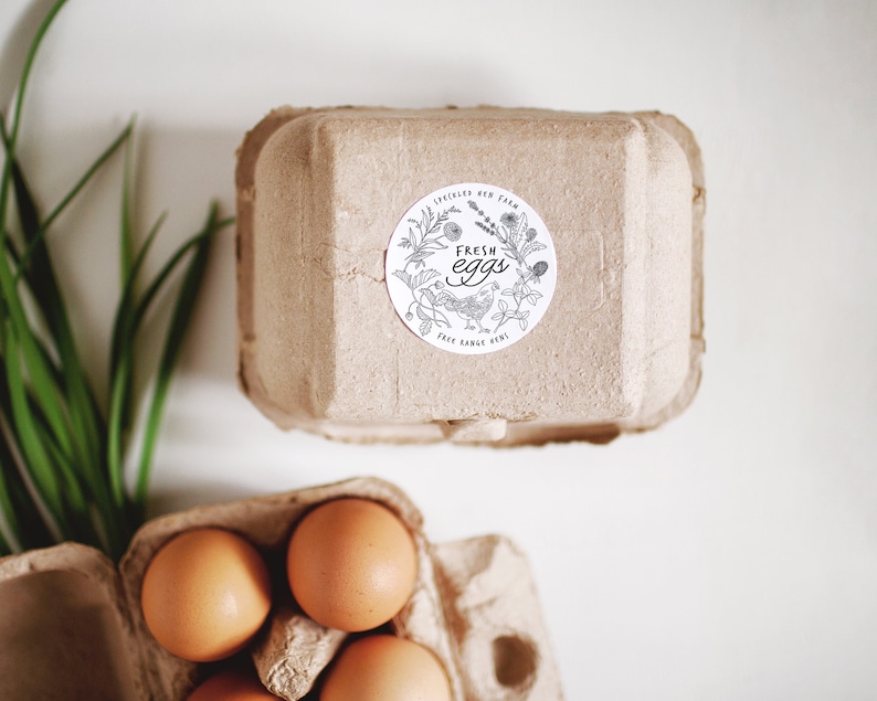 Op de afbeelding: Een kartonnen eierdoos met een sticker waarop "Fresh Eggs" staat met een tekening van een kip en bloemen. De doos is gevuld met bruine eieren.