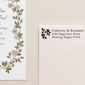 May include: A wedding invitation and envelope set. The invitation features a floral wreath with pink and yellow roses. The envelope is cream-colored with a black floral design and the names Catherine & Benjamin and address information.