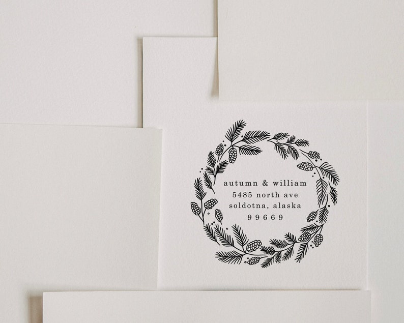 Peut inclure: Illustration en noir et blanc d'une couronne de branches de pin et de pommes de pin. Le texte "autumn & william 5485 north ave soldotna, alaska 99669" est &agrave; l'int&eacute;rieur de la couronne.