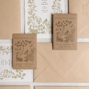 K&ouml;nnte beinhalten: Eine Sammlung von Schreibwaren, darunter Einladungen, Sament&uuml;ten und Umschl&auml;ge. Die Einladungen zeigen botanische Illustrationen und Text, einschlie&szlig;lich Babynamen und Geburtsdaten. Die Sament&uuml;ten sind mit "Wildflower Seeds" beschriftet.