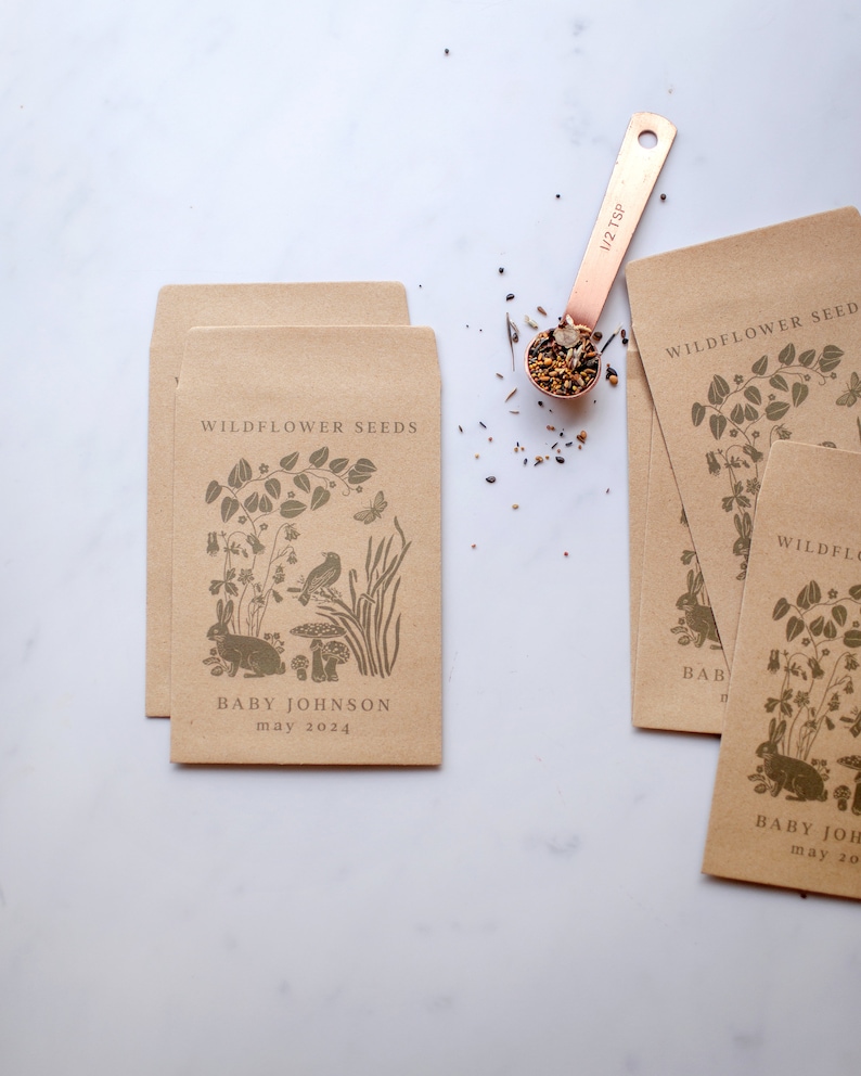 K&ouml;nnte beinhalten: Mehrere Saatgutt&uuml;ten aus Kraftpapier mit einer botanischen Illustration und dem Text "WILDFLOWER SEEDS". Die T&uuml;ten zeigen ein Kaninchen-, Vogel- und Pilzdesign. Der Text "BABY JOHNSON may 2024" ist auf den T&uuml;ten aufgedruckt. Ein Messl&ouml;ffel aus Kupfer enth&auml;lt Samen.