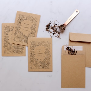 Op de afbeelding: Kraftpapieren zaadzakjes met zwarte bloemenillustraties en de namen "Emma & Robert" en de datum "05.26.24". Een koperen maatlepel bevat zaden, met wat gemorst in de buurt. Twee extra enveloppen zijn ook zichtbaar.