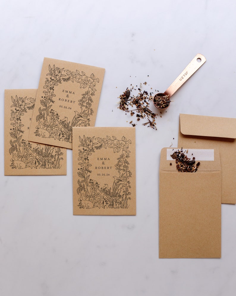 Op de afbeelding: Kraftpapieren zaadzakjes met zwarte bloemenillustraties en de namen "Emma & Robert" en de datum "05.26.24". Een koperen maatlepel bevat zaden, met wat gemorst in de buurt. Twee extra enveloppen zijn ook zichtbaar.