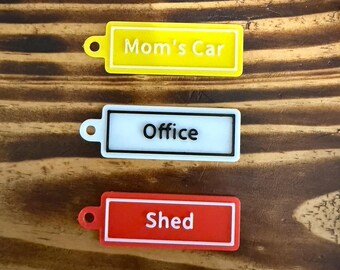 May include: Three rectangular keychains with text. The top keychain is yellow with the text "Mom's Car". The middle keychain is white with black text that reads "Office". The bottom keychain is red with the text "Shed".