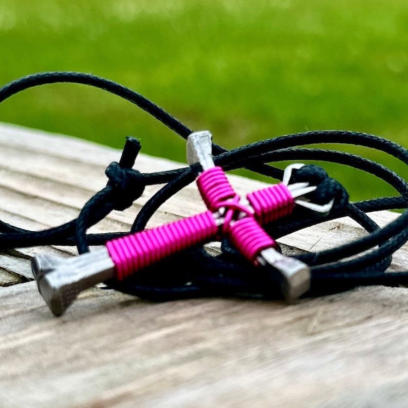May include: A pink wire-wrapped horseshoe nail cross pendant on a black cord necklace.