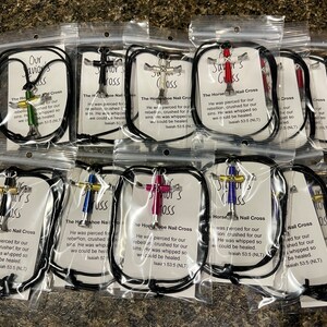 May include: A collection of ten small crosses made from nails, each with a different coloured nail head. The crosses are displayed in individual clear plastic bags with black cords and a small card with the text "Our Saviour's Cross" and a Bible verse about Jesus' sacrifice.