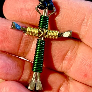 May include: A handmade cross pendant crafted from recycled nails, wrapped with green and gold wire. The pendant is attached to a black cord.