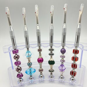 May include: A collection of six seam rippers with decorative beaded handles in various colors, including purple, blue, pink, green, and red. Each ripper has a silver metal blade and handle, displayed in a clear plastic holder.