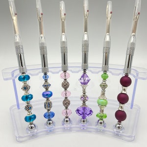 May include: Six silver tools with clear plastic covers and colorful beaded handles. The tools are displayed on a clear plastic stand.