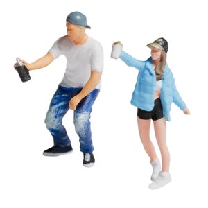 Puede incluir: Dos figuras en miniatura, una con una camisa blanca, pantalones vaqueros azules y una gorra de béisbol azul, sosteniendo una lata de aerosol, y la otra con una chaqueta azul, pantalones cortos negros y una gorra de béisbol negra y blanca, sosteniendo una lata.