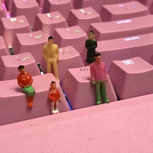 以下が含まれることがあります： ピンクのキーボードの上に立っている人のミニチュア人形が5体。人形はピンク、グリーン、レッド、イエローなど、さまざまな色の服を着ています。