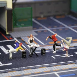 Puede incluir: Escena callejera con figuras en miniatura de bailarines de breakdance y un DJ. Las figuras visten atuendos coloridos, uno de ellos haciendo el pino. Un DJ está en una mesa con altavoces. La escena está ambientada en una superficie gris y azul.