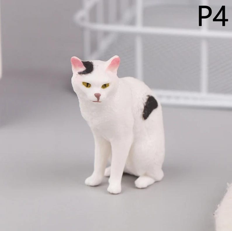May include: A white cat figurine with black spots and pink-tipped ears. The cat has yellow eyes and is sitting upright. The figurine is a decorative item, possibly for a shelf or display.