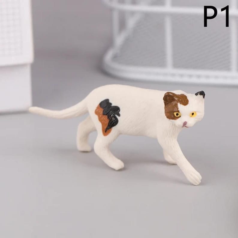 May include: A white cat figurine with brown and black markings. The cat has yellow eyes and is in a walking pose. The figurine has the text "P1" in the upper right corner.