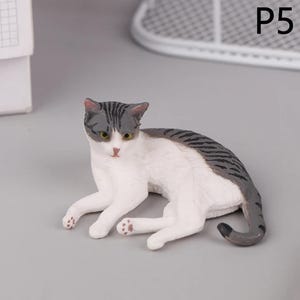 May include: A small, detailed figurine of a cat in a resting pose. The cat has a white body with gray tabby markings on its head and back. It has yellow eyes and pink paws. The figurine is on a gray surface.