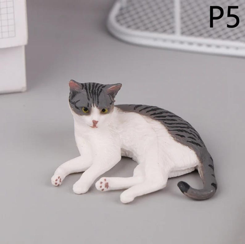 May include: A small, detailed figurine of a cat in a resting pose. The cat has a white body with gray tabby markings on its head and back. It has yellow eyes and pink paws. The figurine is on a gray surface.