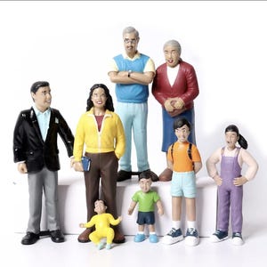 Può includere: Una collezione di figurine di famiglia in miniatura. Le figure includono un uomo in blazer nero, una donna in giacca gialla e bambini in vari abiti. I membri della famiglia sono in piedi insieme, alcuni in abiti casual.